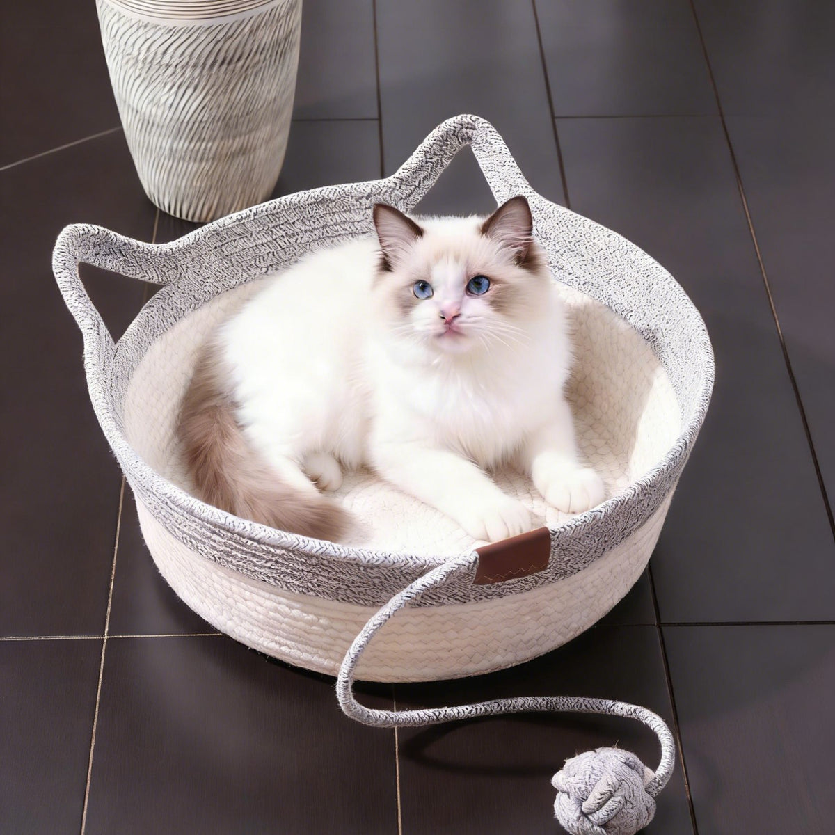 A fluffy white and brown cat with blue eyes sits inside a round woven cat bed with cat-ear handles, placed on a dark tiled floor.